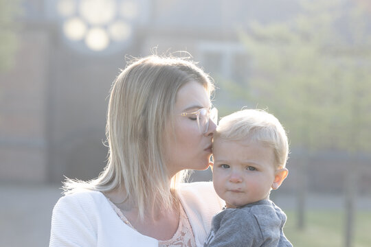 Happy young mother with a small child or todler in her arms, gently kissing his head, standing in the middle of a park in the rays of the rising morning sun. High quality photo