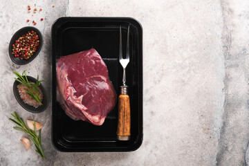 Beef steak. Fresh raw beef steak with fork and rosemary in vacuum black plastic pack on grey stone background. Vacoom package. Top view. Mockup for design idea.