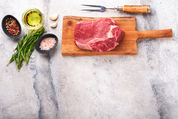 Beef steak. Fresh raw beef rib eye steak with fork rosemary, salt and pepper on old wooden stand on grey stone background. Top view. Mockup for design idea.