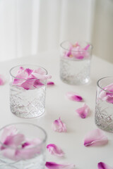 crystal glasses with tonic and natural floral petals on white tabletop and blurred background.