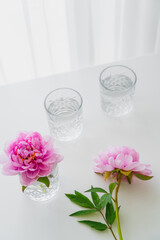 high angle view of faceted glasses with water and pink peonies on white surface.