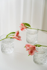 crystal glasses with water near alstroemeria flowers on white tabletop and grey background.
