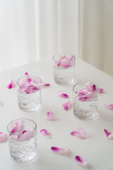 faceted glasses with gin tonic near floral petals scattered on white tabletop on grey background.