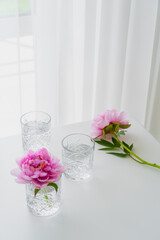 transparent faceted glasses and pink peonies on white tabletop near curtain.