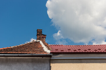Old chimney made of bricks on a roof with ceramic tiles with a cloud above the home next to a house with new metallic rooftop. Construction backdrop