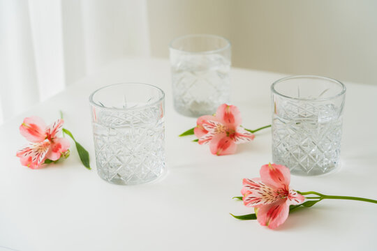 crystal glasses with water near pink flowers on white surface and grey background.
