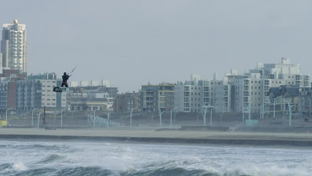 A kiteboarder jumps a wave and does a trick high in the air; in the background are the buildings of the city of Scheveningen, The Netherlands.