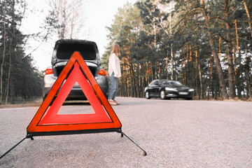 Red emergency triangle with blurred car and woman calling car mechanic in the background. Accident and broken car on the road