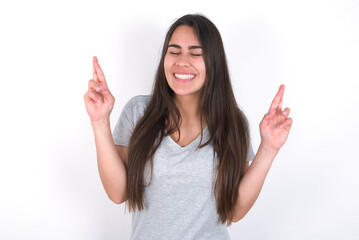 Young beautiful brunette woman wearing grey T-shirt over white wall has big hope, crosses fingers, believes in good fortune, smiles broadly. People and wish concept