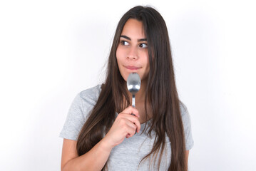 Very hungry Young beautiful brunette woman wearing grey T-shirt over white wall holding spoon into...