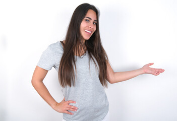 Young beautiful brunette woman wearing grey T-shirt over white wall feeling happy and cheerful, smiling and welcoming you, inviting you in with a friendly gesture