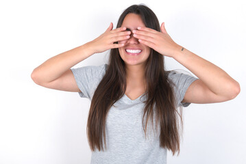 Fototapeta premium Young beautiful brunette woman wearing grey T-shirt over white wall covering eyes with hands smiling cheerful and funny. Blind concept.