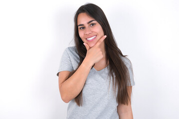 Fototapeta premium Young beautiful brunette woman wearing grey T-shirt over white wall looking confident at the camera smiling with crossed arms and hand raised on chin. Thinking positive.