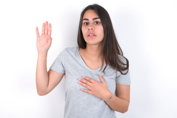 Obraz na płótnie Canvas Young beautiful brunette woman wearing grey T-shirt over white wall Swearing with hand on chest and open palm, making a loyalty promise oath