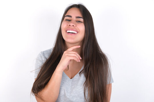 Optimistic Young beautiful brunette woman wearing grey T-shirt over white wall keeps hands partly crossed and hand under chin, looks at camera with pleasure. Happy emotions concept.