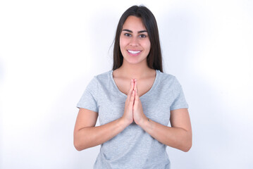 young beautiful brunette woman wearing grey t-shirt over white wall praying with hands together asking for forgiveness smiling confident.