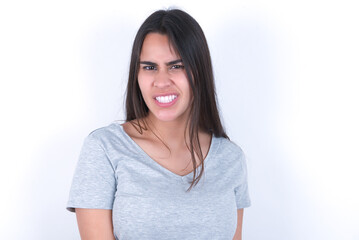 Obraz premium Young beautiful brunette woman wearing gray t-shirt over white wall keeps teeth clenched, frowns face in dissatisfaction, irritated because of much duties.