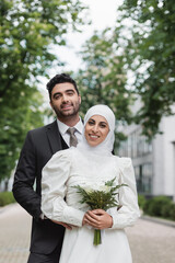 cheerful muslim bride in wedding hijab and white dress holding bouquet near groom outside.