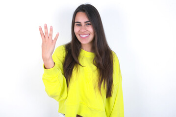 Young beautiful brunette woman wearing yellow hoodie over white wall smiling and looking friendly, showing number four or fourth with hand forward, counting down