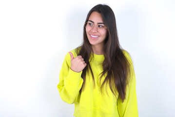 Young beautiful brunette woman wearing yellow hoodie over white wall points away and gives advice demonstrates advertisement
