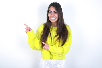 Young beautiful brunette woman wearing yellow hoodie over white wall points at copy space indicates for advertising gives right direction