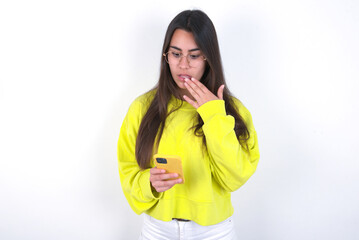 Young beautiful brunette woman wearing yellow hoodie over white wall being deeply surprised, stares...