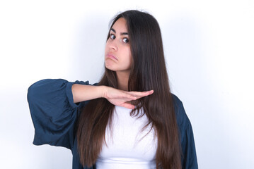 Young beautiful brunette woman wearing blue jacket over white wall, cutting throat with hand as...