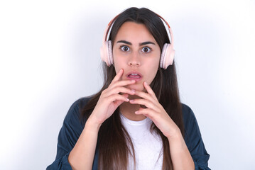 Shocked Young beautiful brunette woman wearing blue jacket over white wall, stares fearful at camera keeps mouth widely opened wears wireless stereo headphones on ears