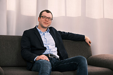 Portrait of young caucasian smiling businessman wearing jacket and glasses sitting on sofa in an open position looking at the camera against background of large window with curtain. Concept of success