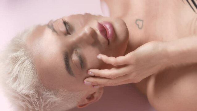 Good-looking young slim Caucasian woman with short light hair turns her head to the camera, opens her eyes and strokes her face through rippling water on pale pink background | Moisturizing commercial