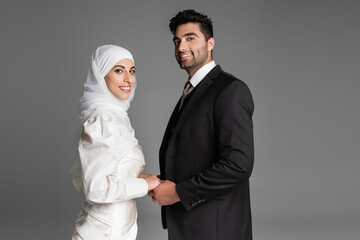 happy muslim bride in wedding dress and groom in suit holding hands isolated on grey.