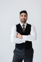 happy muslim man in vest standing with crossed arms isolated on grey.