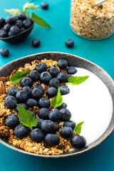 Homemade granola with yogurt, blueberries and mint leaves in a bowl on a turquoise background, delicious healthy breakfast