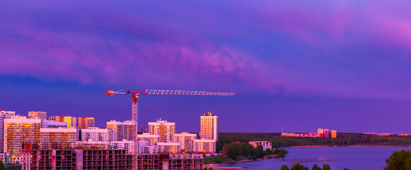 Multi-storey buildings illuminated by the setting sun. Panorama of the evening city with a construction site and a crane against the backdrop of a sunset sky. Beautiful view of the modern city.