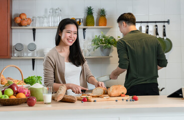 Selective focus of young Asian woman standing in the kitchen, happy beautiful wife cutting bread and handsome husband helping her cooking and prepare food at background at home
