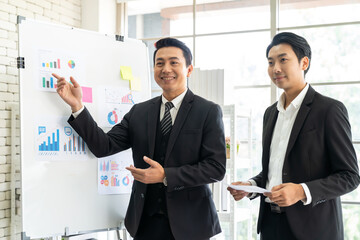Two Asian business man present business strategy plan to corporate colleagues in the office. Group of Asian businesspeopole present business plan on white board to his parther in the modern office.