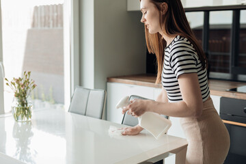 Side view of young pretty woman standing in kitchen, spraying on white table with vase from...