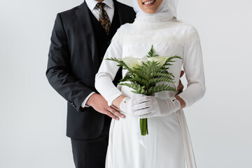 cropped view of groom hugging happy bride in wedding dress with bouquet isolated on grey.