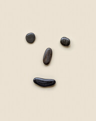 Anthropomorphic face from black colored sea stones on beige background, cheerful, happy, joyful mood, smile on the face. Summer creative flat lay, minimal style, top view