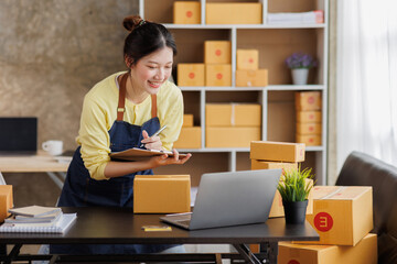 Obraz na płótnie Canvas Startup SME small business entrepreneur of freelance Asian woman wearing apron using laptop and box to receive and review orders online to prepare to pack sell to customers, online sme business ideas.
