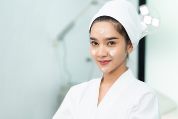 Beautiful Asian woman in bathrobe and towel on head with facial treatment cream applied on her face, smiling looking at camera while doing a beauty treatments procedures in a clinic treatment room.