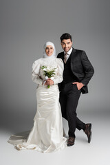 happy groom in suit posing near muslim bride with wedding bouquet on grey.