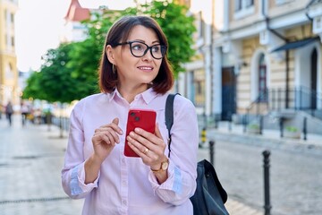Middle aged smiling woman looking into smartphone on city street