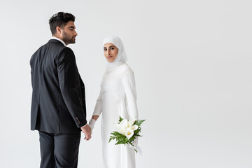 smiling muslim bride in wedding dress with bouquet of calla lily holding hands with groom isolated on grey.