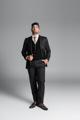 full length of stylish muslim groom in suit posing with hand in pocket while standing on grey.