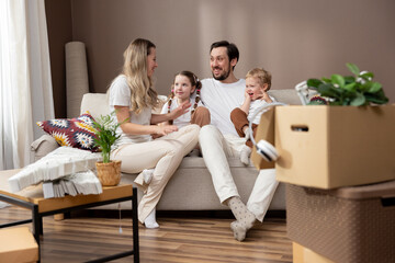 Loving family spends time with each other during move relaxing on couch, talking, discussing. Parents with two children enjoy their new apartment, cardboard boxes with unpacked belongings all around.