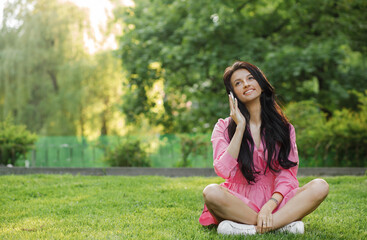 Young happy woman talking on cell phone sitting on grass in summer city park.