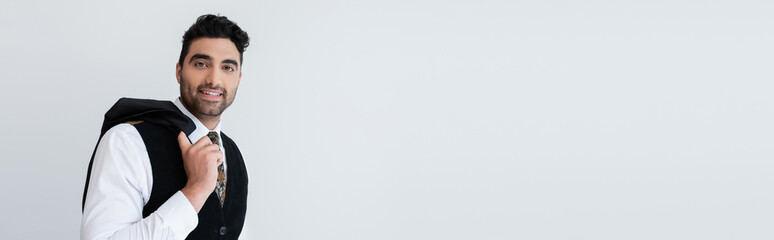 happy muslim man in vest standing and holding blazer isolated on grey, banner.