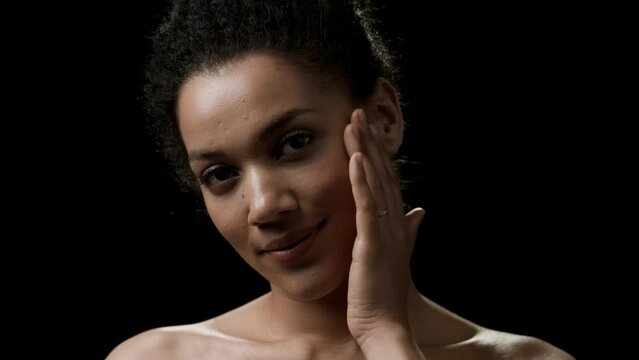 Beautiful slim African American young black-haired woman strokes her face looking at the camera smiling on black background | Sagging jowls prevention concept