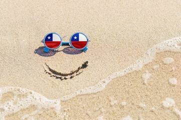 Fototapeta na wymiar A painted smile on the beach and sunglasses with the flag of the Chile. The concept of a positive holiday in the resort of the Chile.